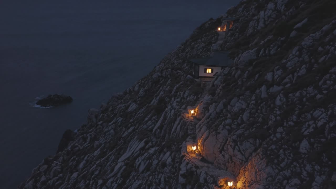 Illuminated cabins perch on a rugged cliffside at dusk, connected by a winding path with glowing lights, creating a serene atmosphere overlooking a tranquil ocean and distant rock formation