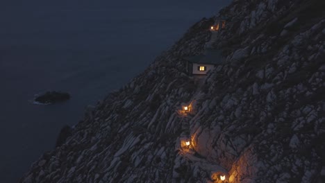 illuminated cabins perch on a rugged cliffside at dusk, connected by a winding path with glowing lights, creating a serene atmosphere overlooking a tranquil ocean and distant rock formation