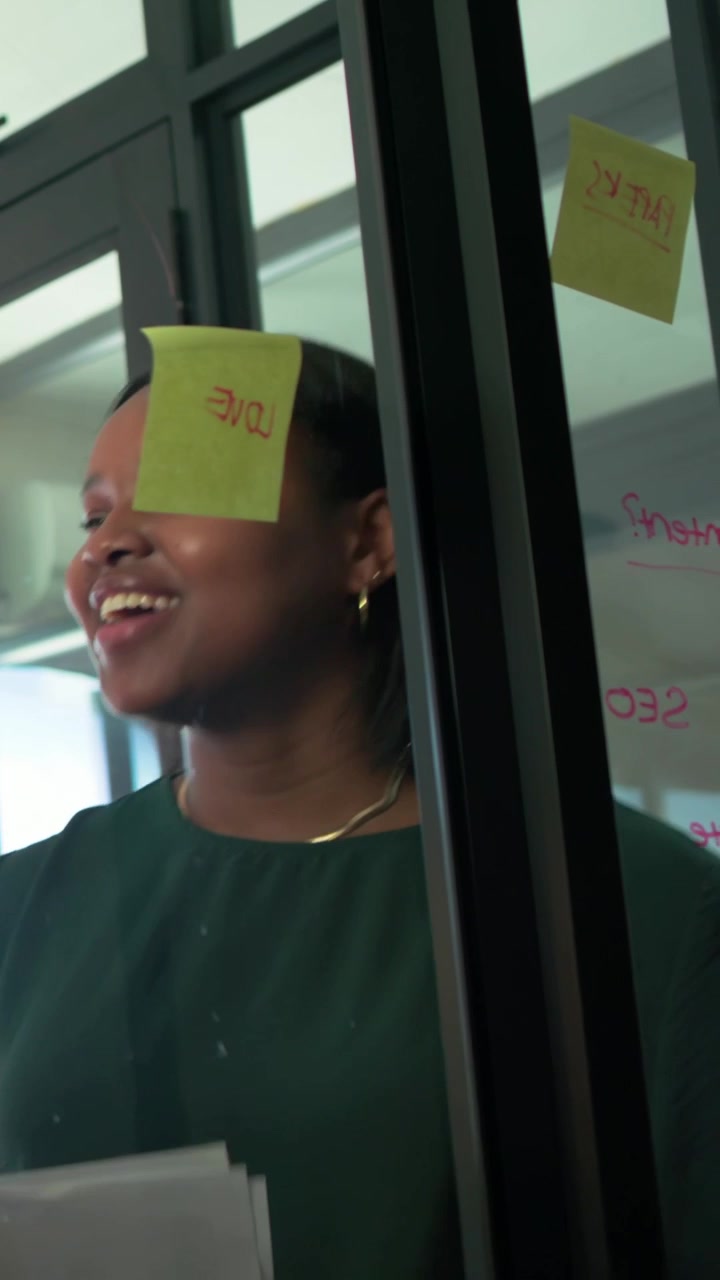 Video verticale: Donna con un top verde che legge documenti e presenta in una stanza di vetro, affiggendo appunti e scrivendo.