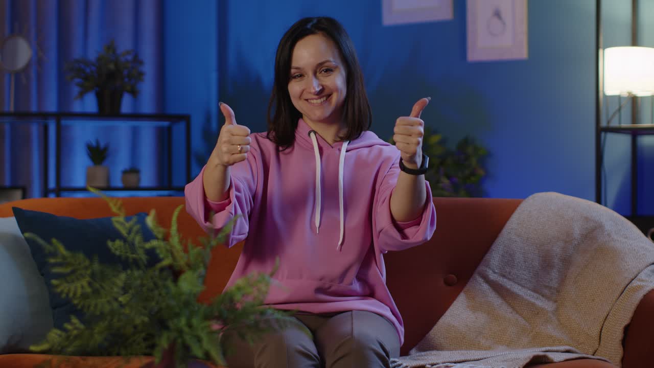 Portrait of happy woman sitting on sofa at home raises thumbs up likes good agrees with positive