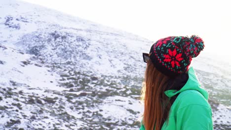 Medium-shot-of-a-woman-in-a-green-jacket-and-a-hat-looking-out-over-a-snowy-landscape-on-a-sunny-day