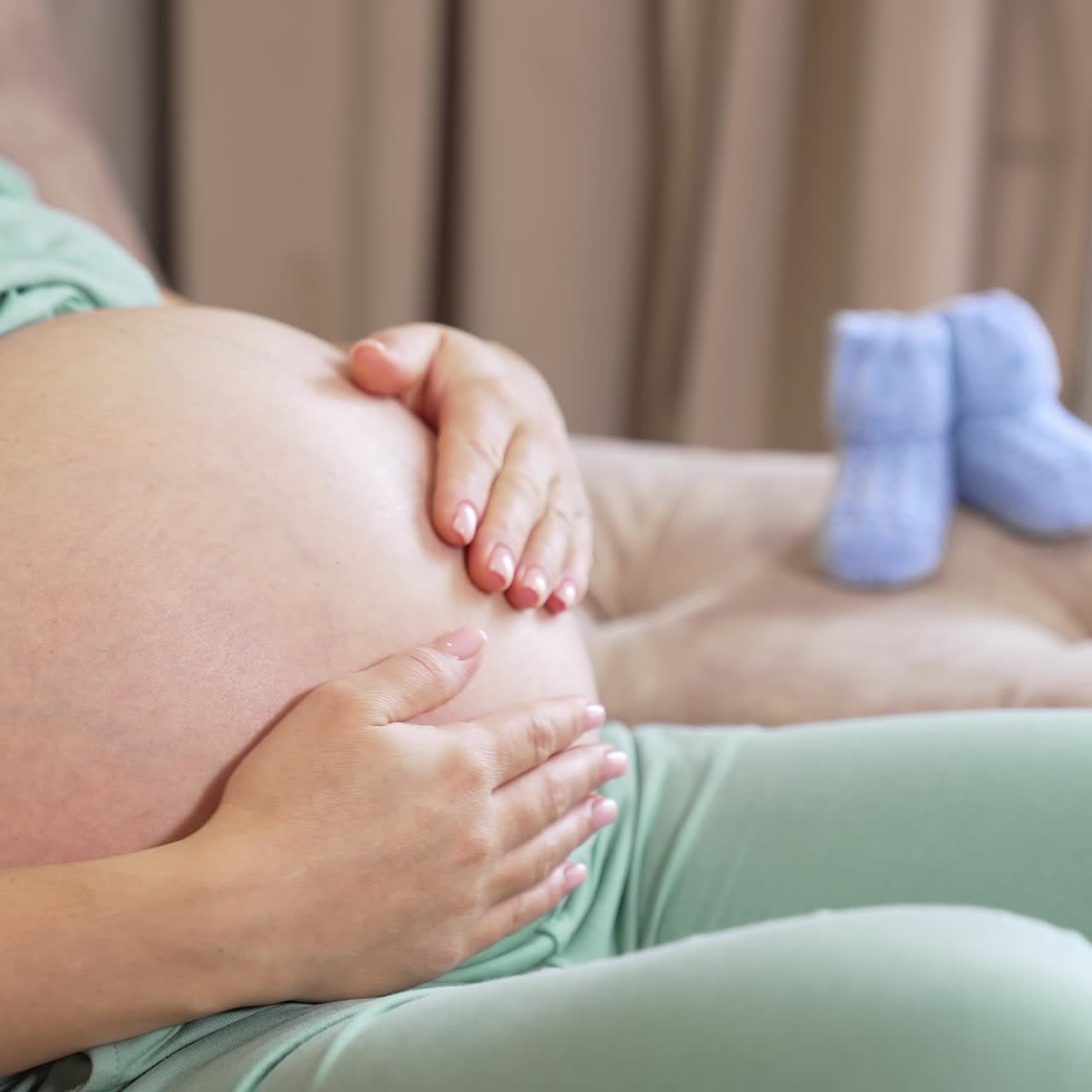 Una futura madre premurosa accarezza la sua grande pancia mentre riposa sul divano. La donna accarezza il suo bambino nella grande pancia. Due piccole scarpine da neonato sono sul bracciolo sullo sfondo.