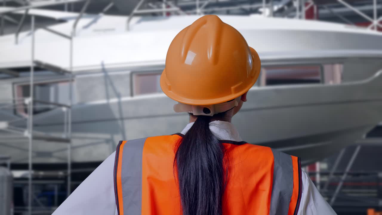 Primo piano da dietro di un'ingegnera con casco di sicurezza che si guarda intorno, in piedi e con le mani sui fianchi, con costruzione di yacht in un moderno cantiere navale