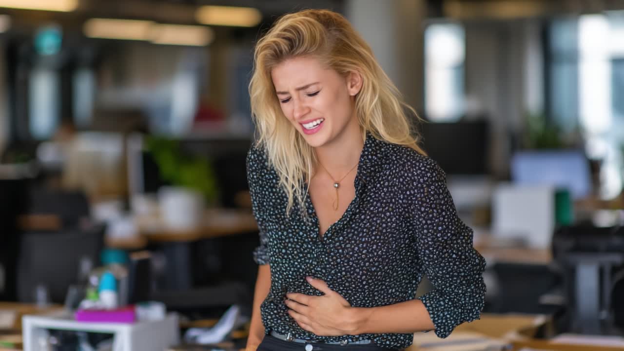 A Young Woman in an Office Struggles with Discomfort, Gripping Her Stomach in Pain, Highlighting the Realities of Workplace Health Challenges