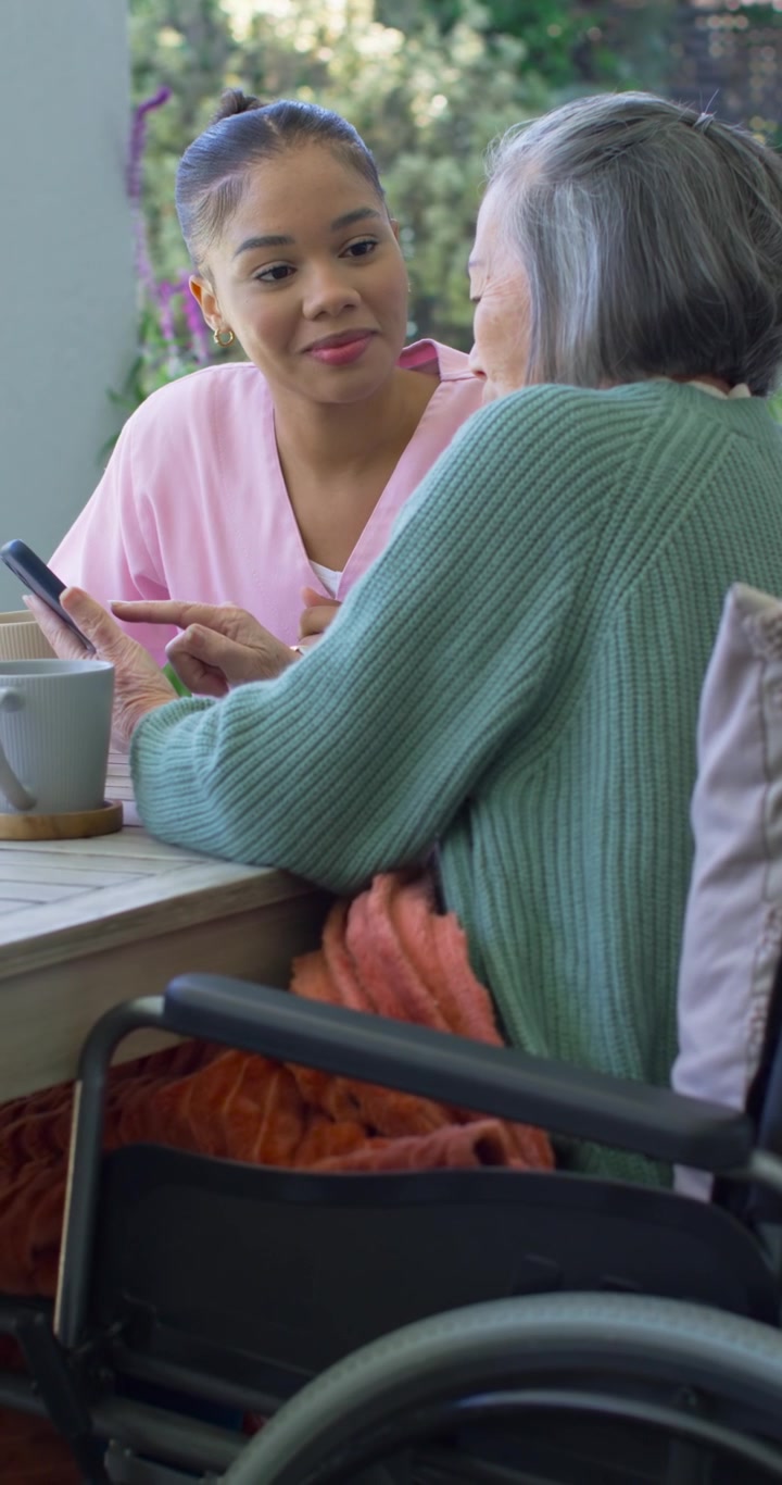 Vertical video: African American woman showing phone, teaching Asian senior in wheelchair on porch