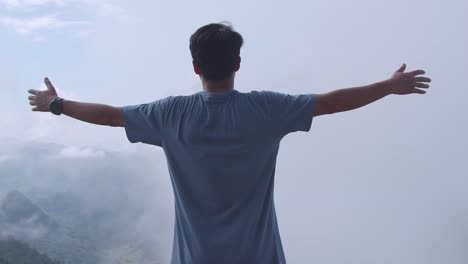 asian hiker male standing on a veranda cliff and raising his hands celebrating reaching up top of foggy mountain