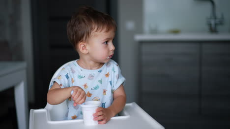 calm quiet baby boy sitting at high chair holding a spoon in a cup and looking aside. lovely toddler eating yogurt.