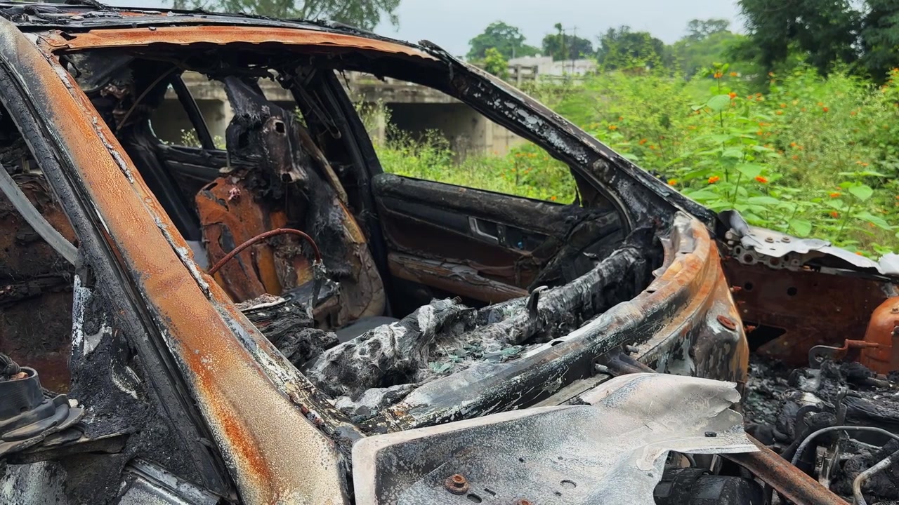 Plano sequência ao longo da área do para-brisa queimado, mostrando um interior de carro carbonizado com a estrutura exposta e danos severos de incêndio.