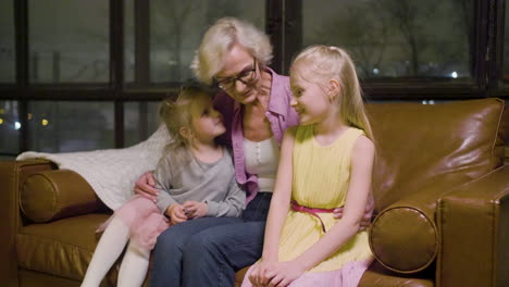 grandmother hugging to her two granddaughters sitting on the sofa while talking in the living room at home