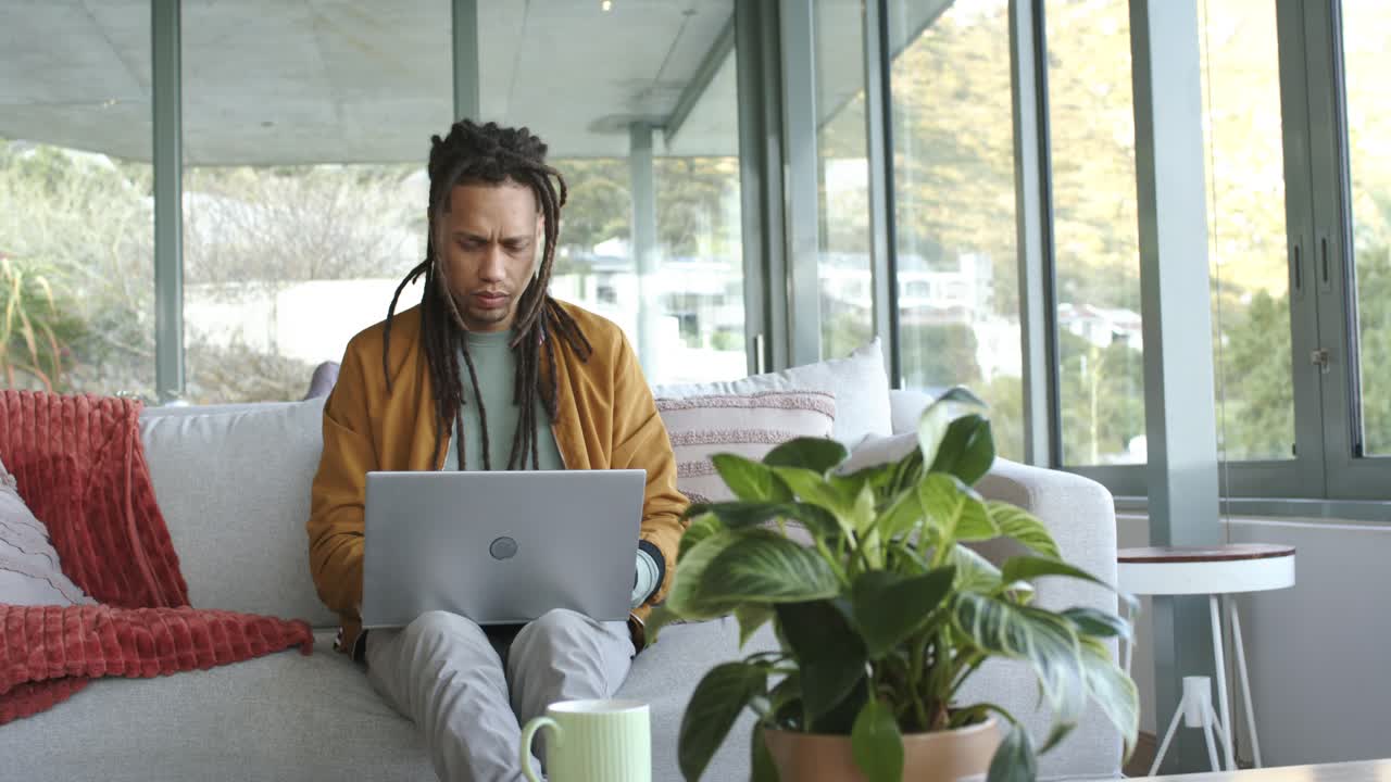 Settling on sofa in living room, man with dreadlocks typing and scrolling on laptop for work