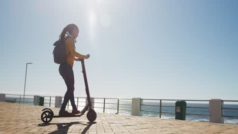 African-american-woman-wearing-headphones-and-backpack-riding-scooter-on-promenade-by-the-sea