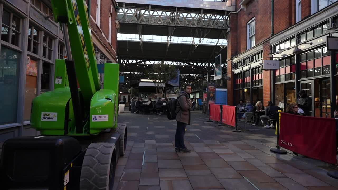 Man walks through Old Spitalfields Market entrance past a green crane and outdoor cafes, London