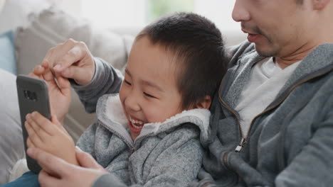 happy asian boy with father using smartphone watching online entertainment on mobile phone touchscreen device browsing internet relaxing on sofa together 4k footage