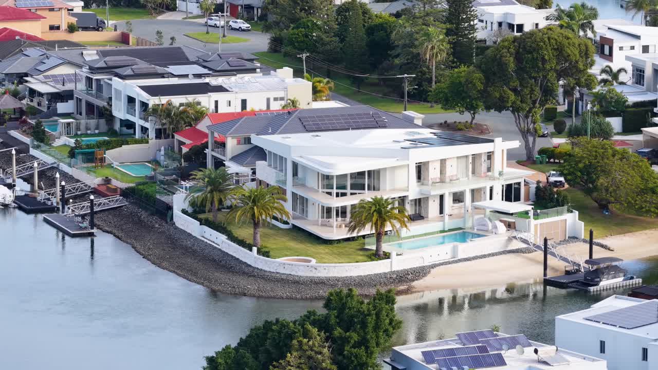 Aerial footage smoothly pans over a modern waterfront mansion with pool, private dock, and lush landscaping in a sunny Gold Coast neighborhood