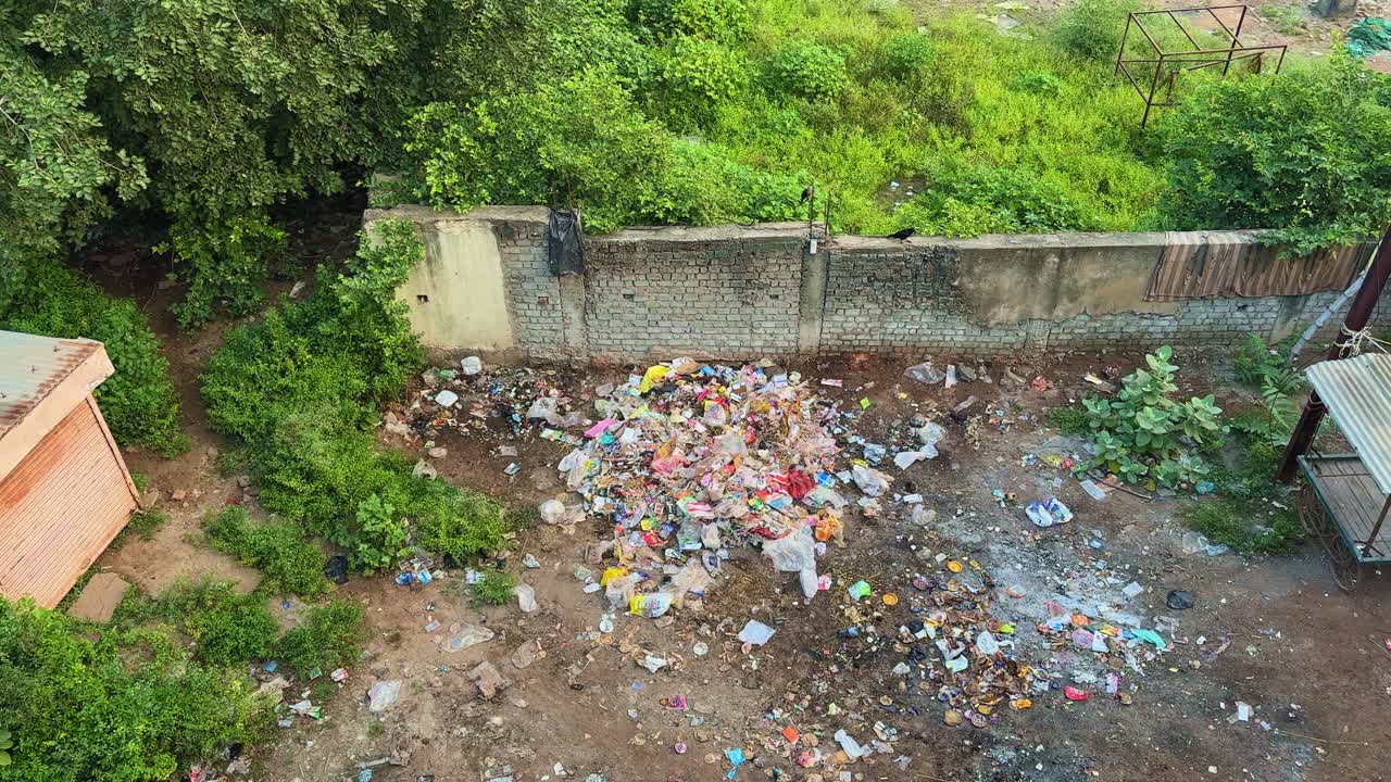 A wide-angle view showing a large pile of garbage near a wall surrounded by greenery and small structures — highlighting pollution amid urban neglect and ecological imbalance