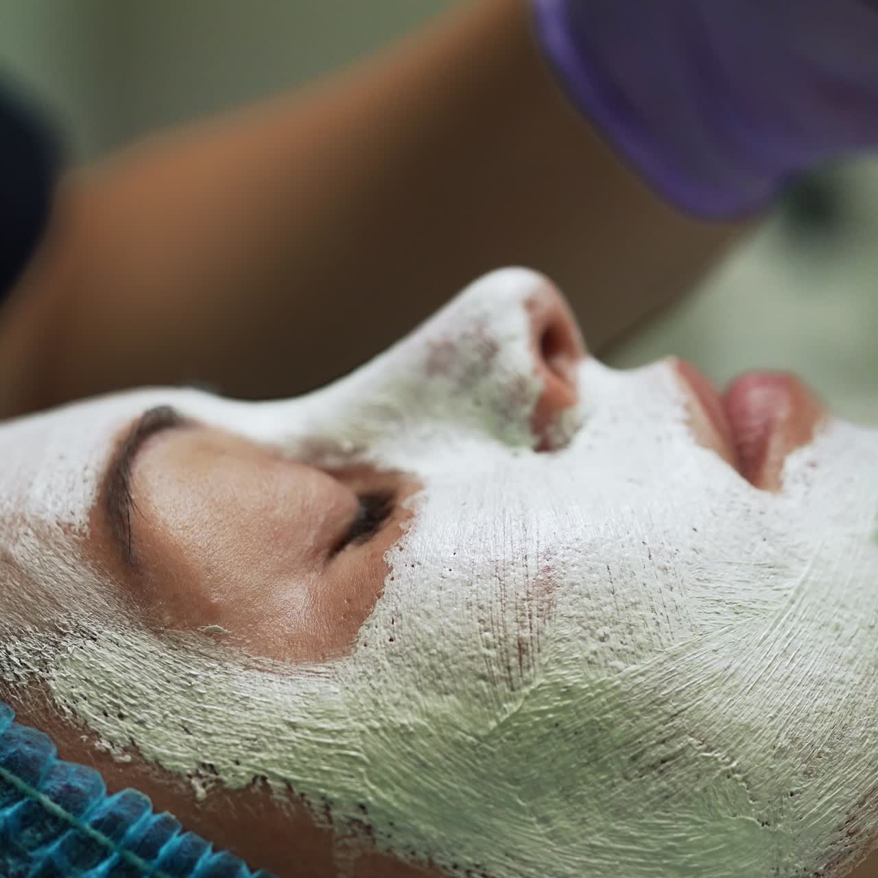 Beautician puts a white mask on the female client's face. Skin care and rejuvenation in modern beauty salon. Side view close up