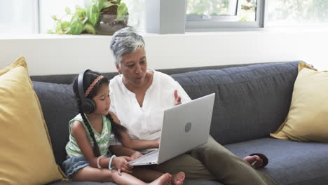 Abuela-Y-Nieta-Birraciales-Sentadas-En-Un-Sofá,-Ocupadas-Con-Una-Computadora-Portátil