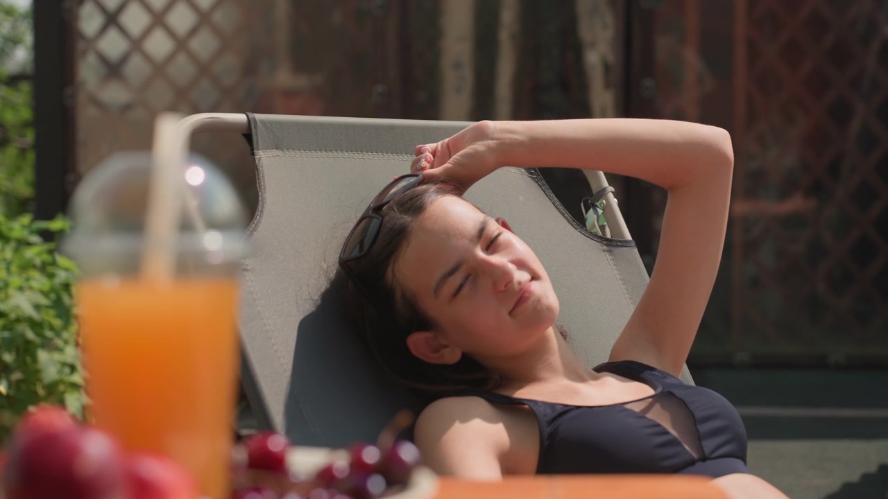 Jovem mulher relaxa em uma espreguiçadeira cinza ao ar livre, usando óculos de sol e ajustando-os com a mão. Um prato de frutas, incluindo cerejas, está colocado por perto em uma mesa de madeira ao lado de uma laranja.