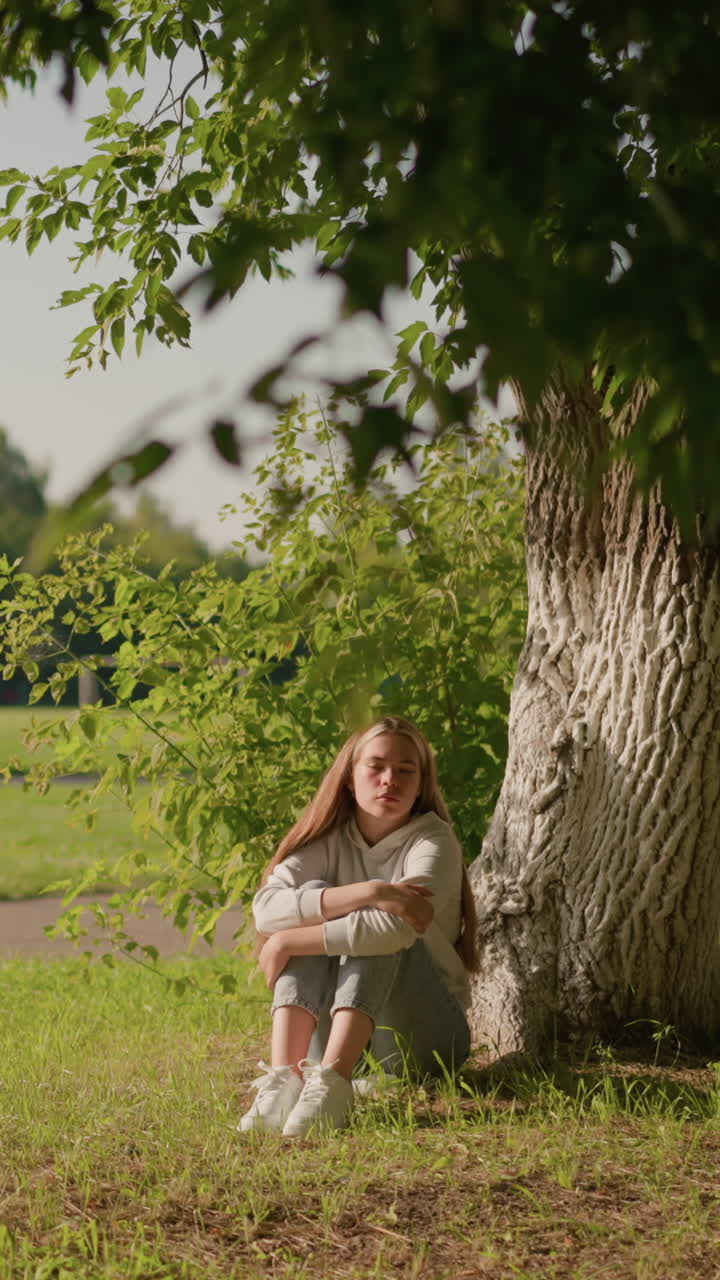 dama se sienta en el suelo cubierto de hierba bajo un árbol con expresión pensativa, brazos cruzados sobre las piernas, la luz del sol se refleja en su cara, el fondo muestra postes y estadio