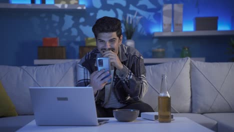 joven en casa por la noche mirando el teléfono y regocijándose.