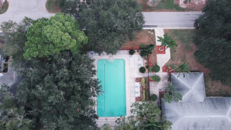 Birdseye-drone-view-over-blue-swimming-pool-next-to-house-with-grey-rood-and-garden-with-large-trees