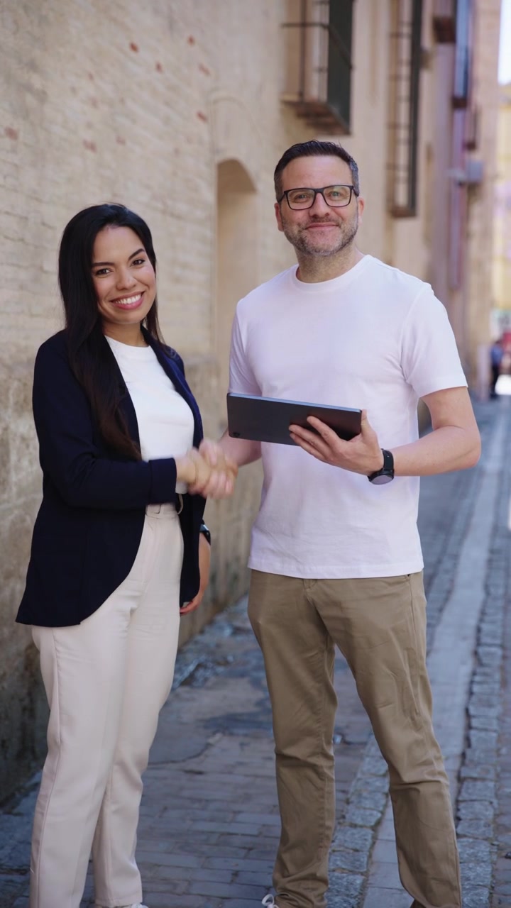 Un empresario y una empresaria con vestimenta casual elegante revisan una tableta digital, chocan las cinco y sonríen a la cámara