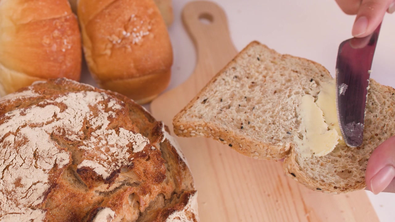 Close-up shot of a knife spreading butter on multigrain bread surrounded by artisanal loaves.