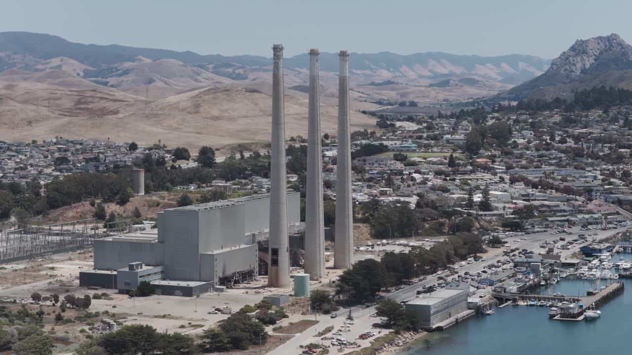 Aerial drone footage of the historic smokestacks from the Morro Bay Power Plant in Morro Bay, California. The coastal landscape, harbor, and nearby cityscape are visible in the background