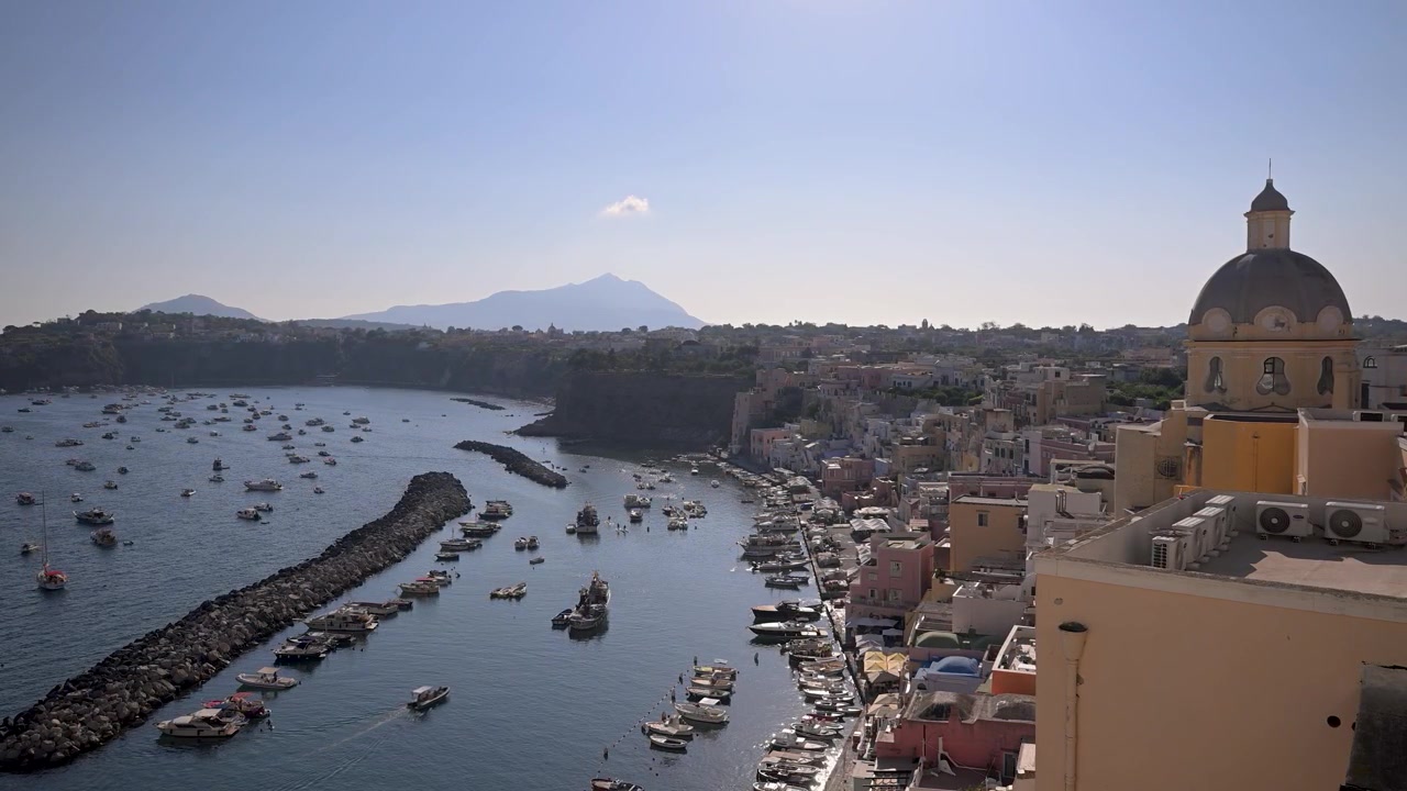 Colorido paseo marítimo de Marina Corricella y Santa Maria delle Grazie Incoronata sobre un puerto concurrido lleno de pequeñas embarcaciones en un día despejado y soleado