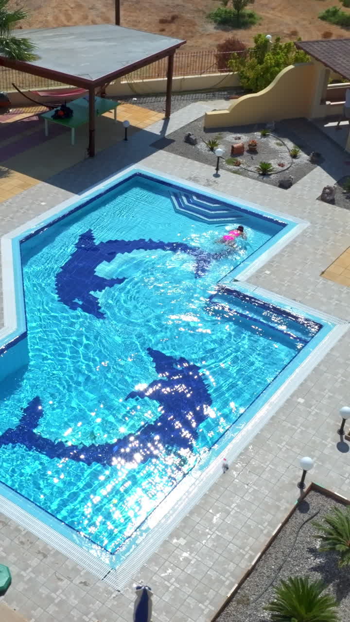 Lodret droneoptagelse omkring en kvinde, der svømmer i en pool i en villa på en solskinsdag