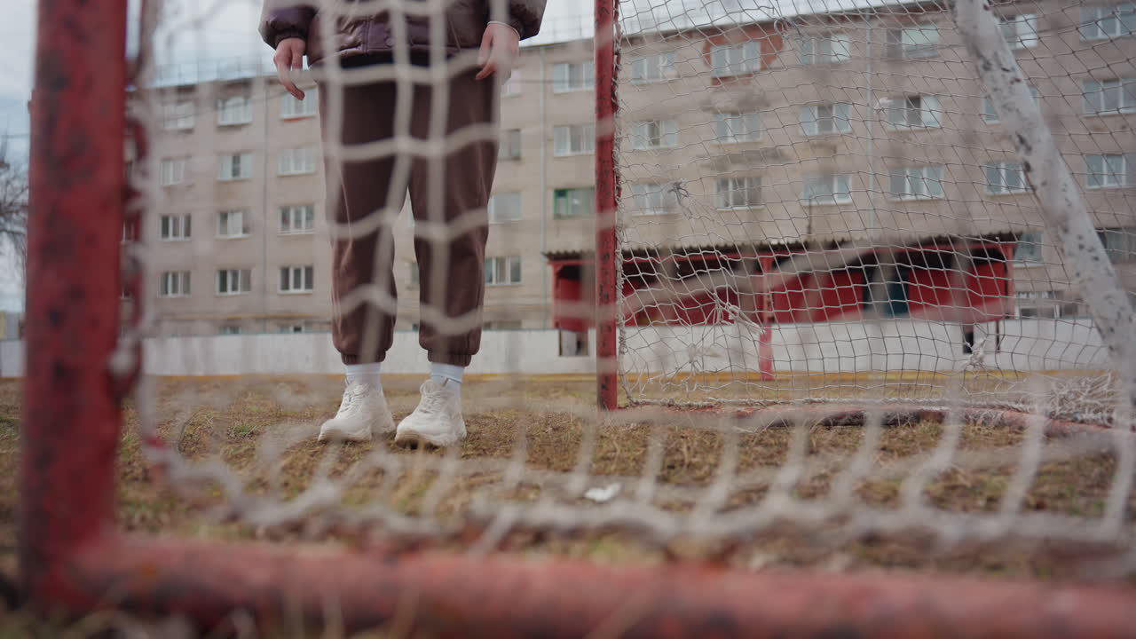 흰머리의 나이 지긋한 여성의 발이 그물망을 뚫고 나아가는 모습, 닳아 해진 잔디와 진흙, 녹슨 붉은 골대 프레임, 아파트 단지 배경이 클로즈업된 골 라인, 질감을 포착하는 솔직한 로우 앵글 구도