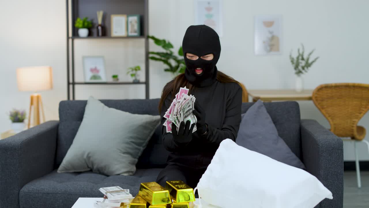 A masked thief counts stacks of money and gold bars on a living room sofa