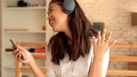 happy young asian woman in earphones singing and listening to music while sitting on bed and holding mobile phone at home