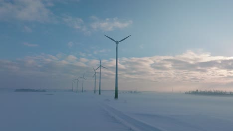 aerial establishing view of wind turbines generating renewable energy in the wind farm, snow filled countryside landscape with fog, sunny winter day, wide ascending drone shot moving forward