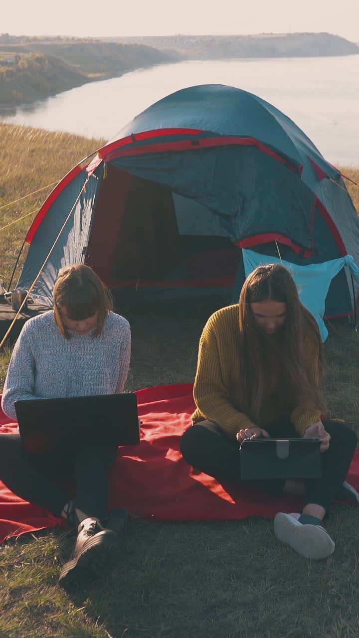 girlfriends work on modern tablet and laptop sitting by blue tent on steep bank against river in windy autumn evening