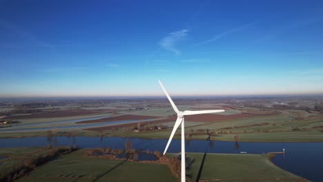 Solar-panels-with-frost-in-front-of-solitary-wind-mill-turbine-part-of-sustainable-industry-in-Dutch-flat-river-landscape-against-blue-sky