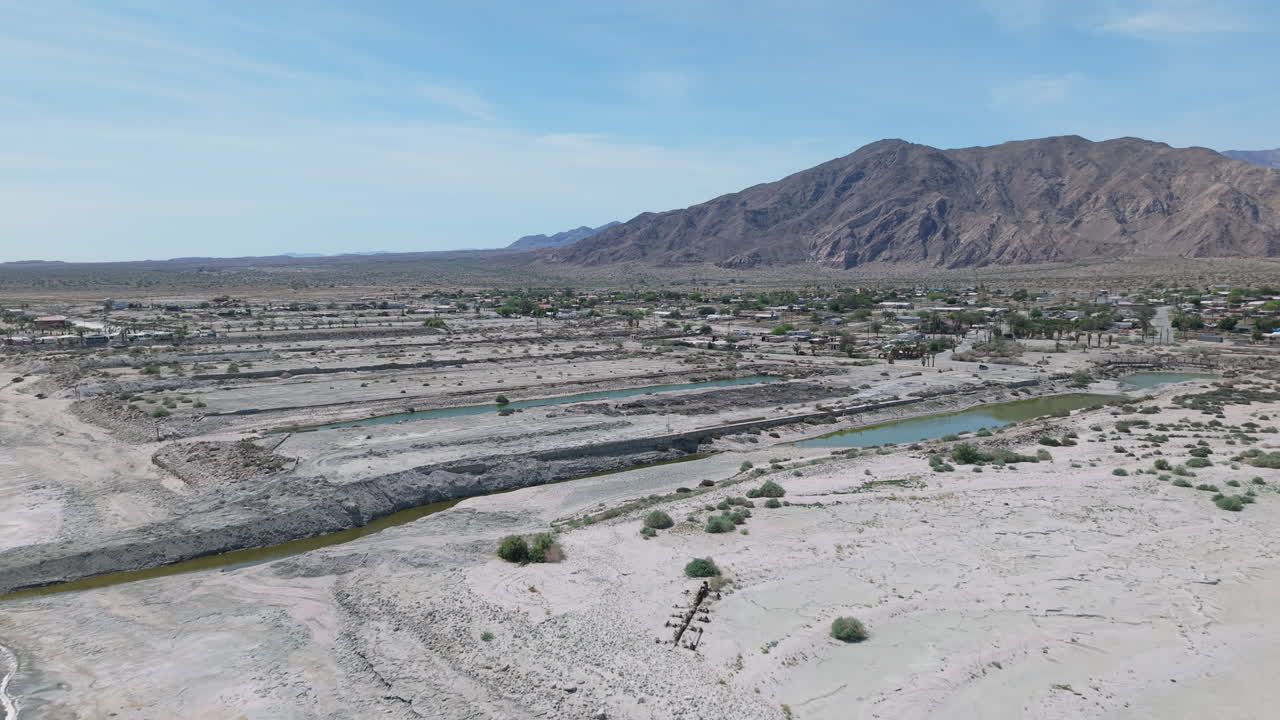 High drone shot flying forward over the Salton Sea toward Desert Shores, California. The retreating shoreline, nearly empty marina basin, and San Jacinto Mountains are visible