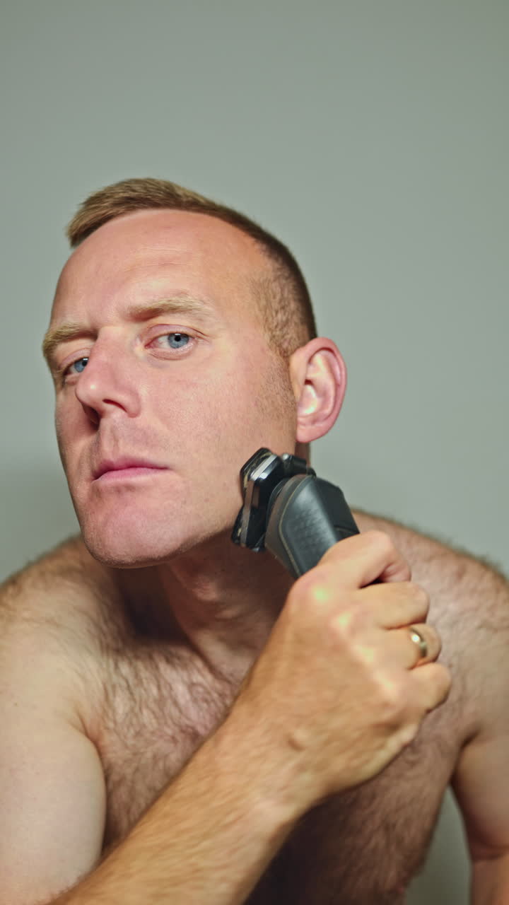 Uomo dagli occhi azzurri che si rade la barba davanti allo specchio di casa. Uomo che si rade con un rasoio elettrico. Igiene del viso, uomo che si prende cura della pelle del viso.