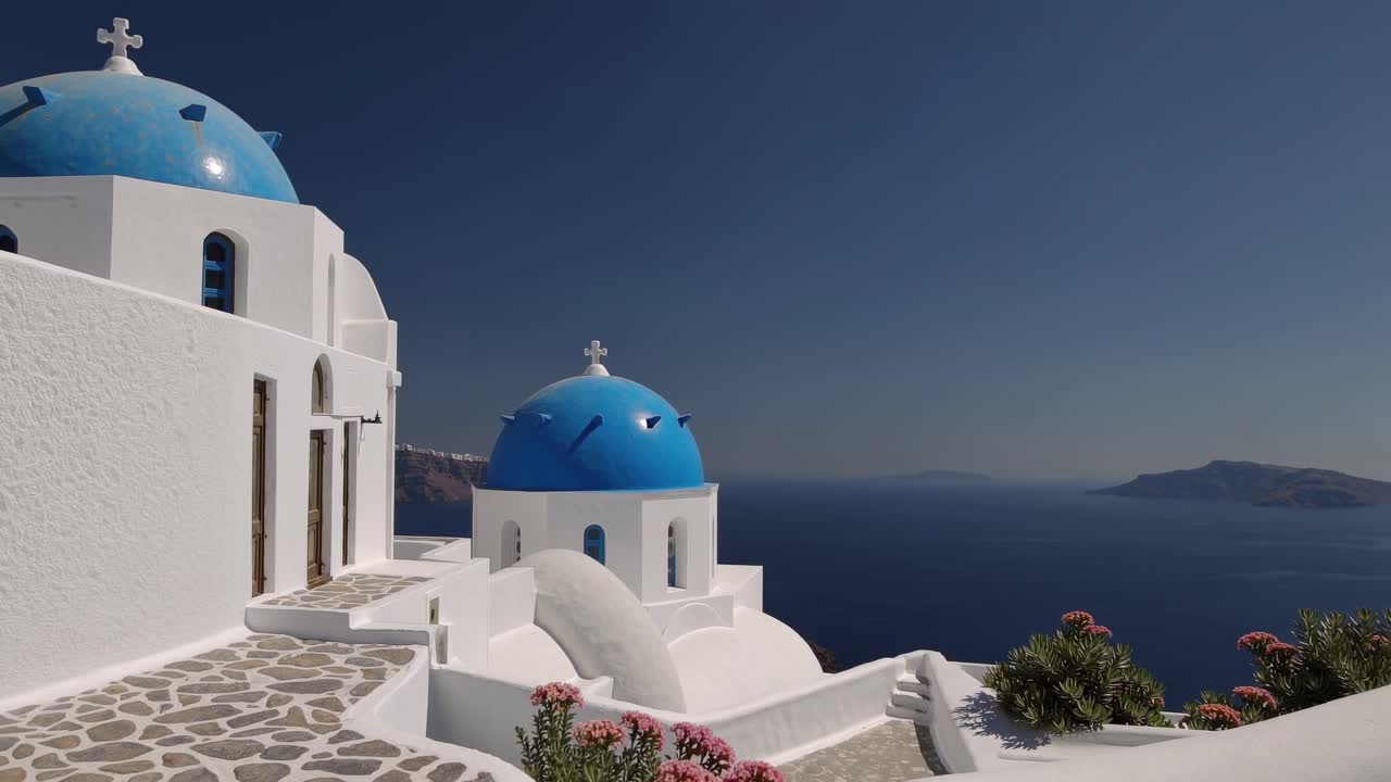 Traditional whitewashed church with iconic blue domes perched on a cliff, overlooking the vast Aegean Sea under a clear blue sky, capturing the essence of Santorini's beauty