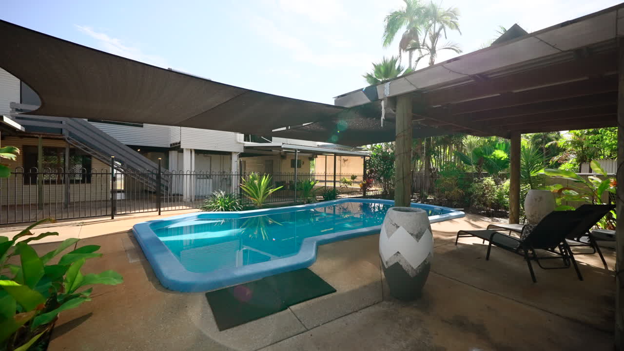 Push through shot of shaded pool on the edge of a large elevated family home. Concrete pool area with sun beds shade sails and gazebo
