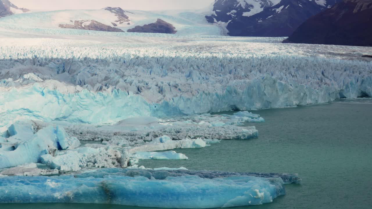 Perito Moreno Glacier with ice formations and fractured front extends into the waters of Lago Argentino near El Calafate. Static shot with mountainous terrain in the background.