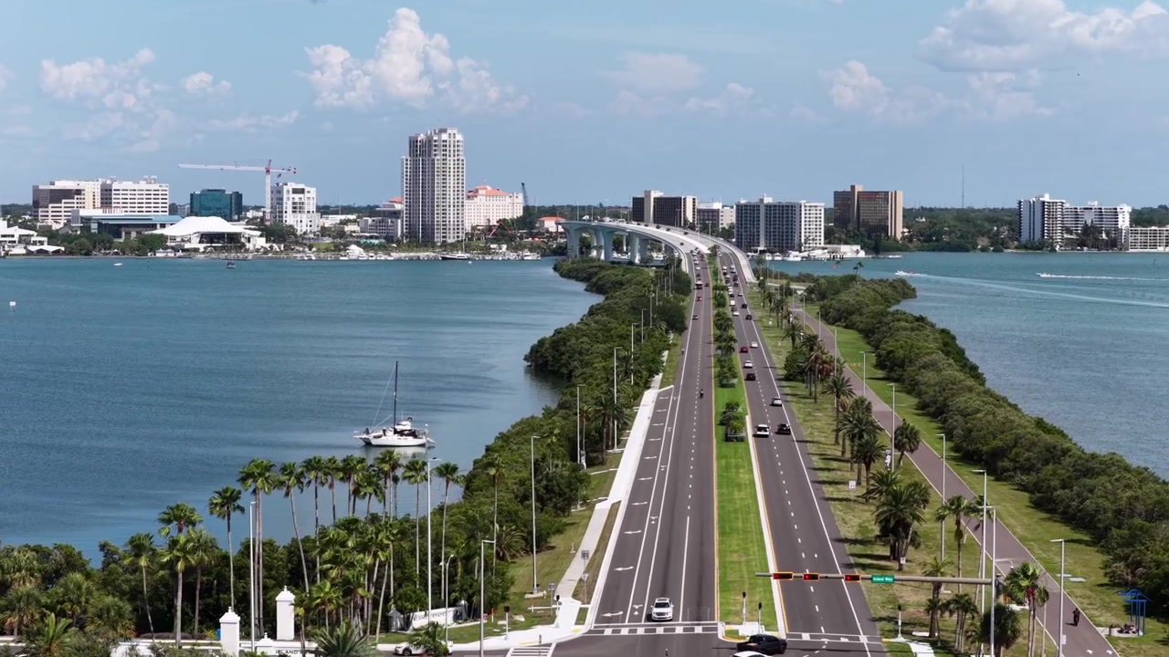 Il ponte rialzato collega Clearwater Beach allo skyline del centro di Clearwater, Florida; una strada fiancheggiata da palme porta il traffico sopra l'Intracoastal Waterway, oltre barche e grattacieli. Ripresa aerea grandangolare con drone.