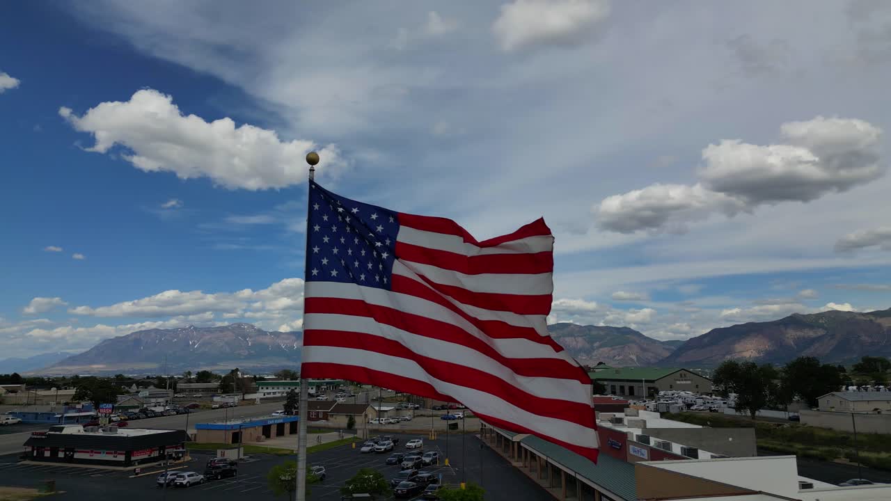 American flag USA blowing waving in the wind on beautiful sunny summer day with clouds and blue skies overlooking mountains and small town America USA as drone flys pans around flagpole - in 4K 60fps