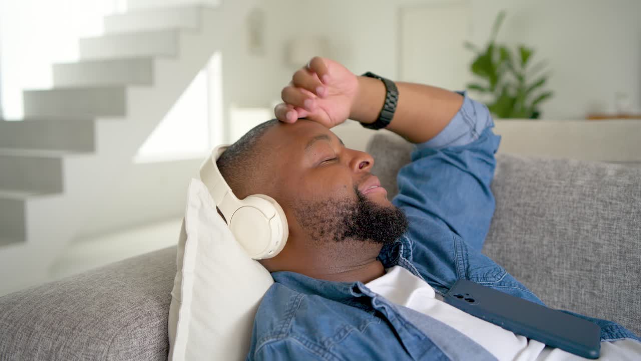 Uomo afroamericano sdraiato sul divano di casa, con il telefono, che ascolta musica in cuffia per rilassarsi, sorridendo
