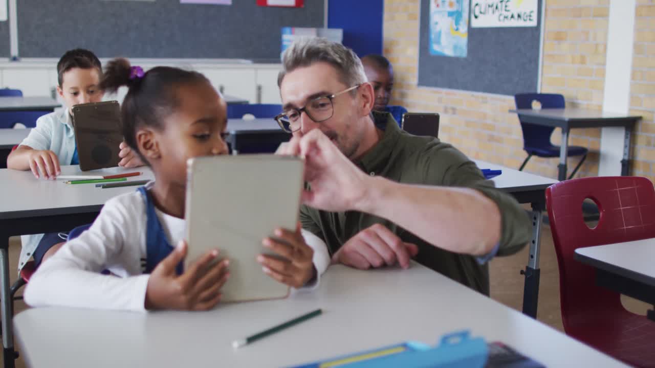 Diverse male teacher helping a schoolgirl sitting in classroom using tablet