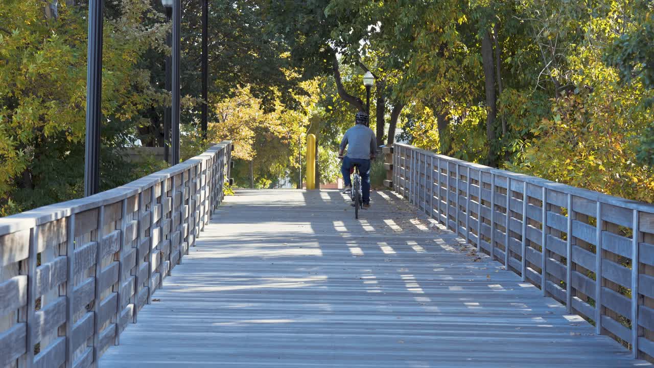 hombre con casco montando bicicleta casualmente en un puente de madera, disfrutando del agradable día soleado, detrás de la vista, filmado en 4k
