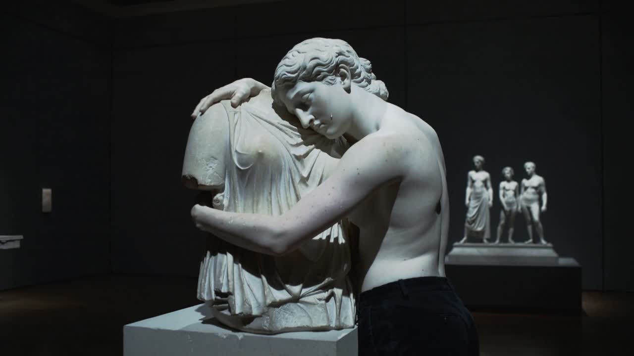 En skjorteløs mann nærmer seg en marmortorso og omfavner den på et museum, med spotlights som avslører skulpturer.