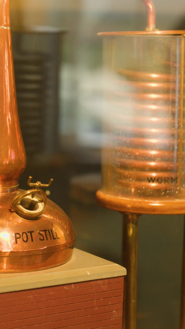 Camera pans across copper pot still, worm tub, and spirit barrels in warm, indoor lighting