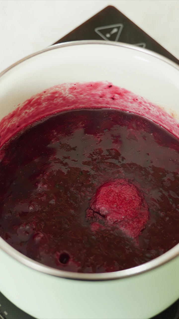 Liquido denso rosso scuro sobbolle in una pentola bianca. Bacche intere galleggiano nella miscela mentre si addensa. La scena è ambientata su uno sfondo di cucina con una messa a fuoco ravvicinata costante.
