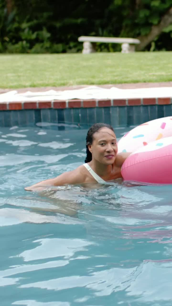 Vertical video: Swimming woman in swimsuit paddling at backyard pool, spotting donut float for rest
